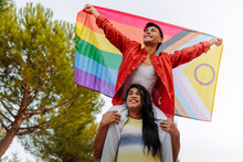 Two happy men, one on another's shoulders, proudly display a vibrant Progress Pride Flag outdoors.