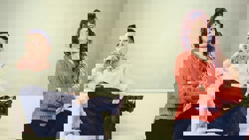 A man and woman sit apart on a couch, appearing upset with each other. The man looks left, while the woman, with crossed arms, glares right.