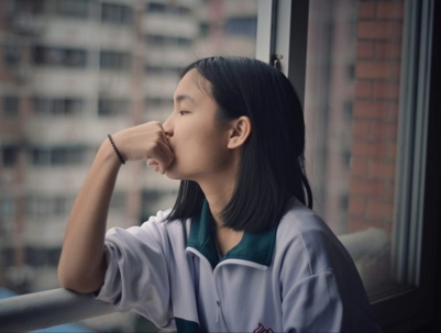 A young woman, eyes closed, looks thoughtfully out a window. She wears a white and green uniform, with blurred buildings in the background.