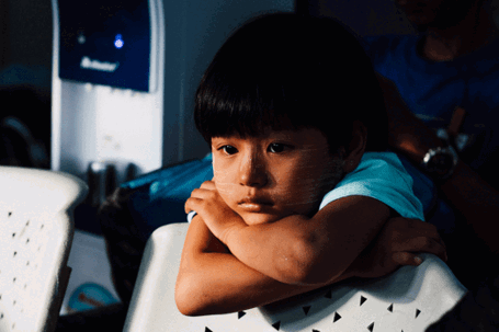 A young child with dark hair sits with their chin resting on crossed arms, looking pensive in what appears to be a waiting room.