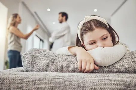 Little girl looking sad on the couch while parents are fighting in the background