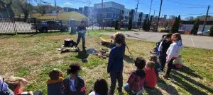 Students gather outdoors for a learning activity. An instructor demonstrates skills around a small fire pit, with buildings in the background.